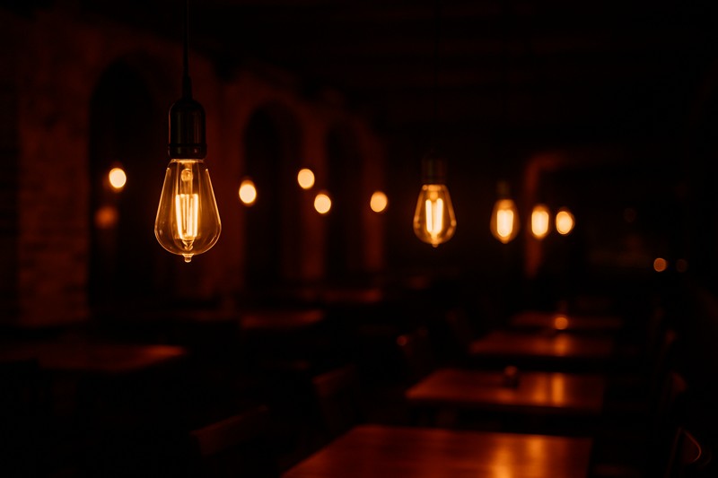 A stunning image of a dimly lit restaurant or bar, with exposed ST64 Edison bulbs with spiral filaments glowing at 2200K. The light is purely atmospheric, creating pools of warm, orange light.