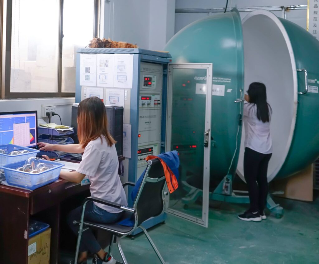 Two technicians testing LED bulbs using a large integrating sphere system, measuring lumen output, CCT, CRI, and other optical performance data in a professional lighting laboratory.
