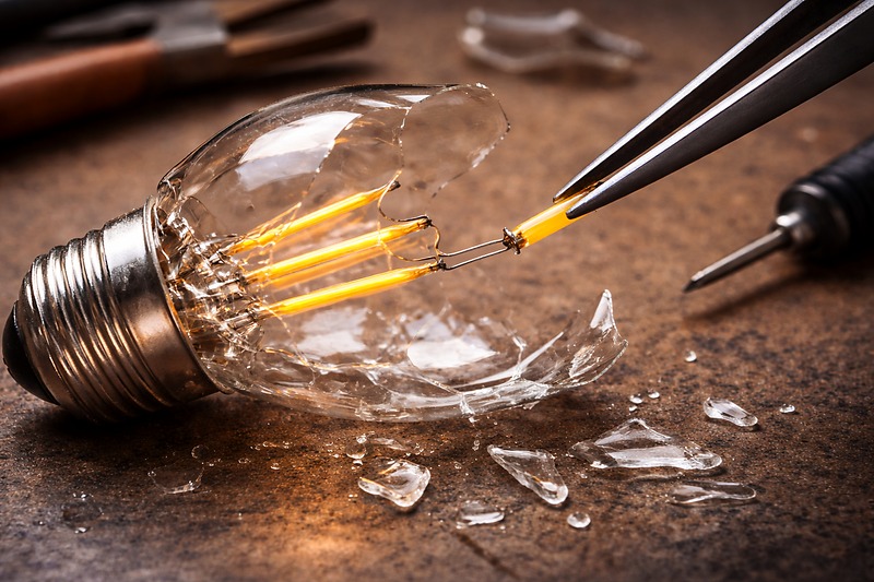 A close-up of a broken LED filament bulb on a workbench. Tweezers are holding a tiny yellow LED filament strand. The glass text is shattered, indicating the difficulty of repair.