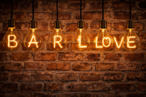 A row of textual light bulbs hanging against a brick wall. The glowing filaments inside spell out the word "BAR" and "LOVE".