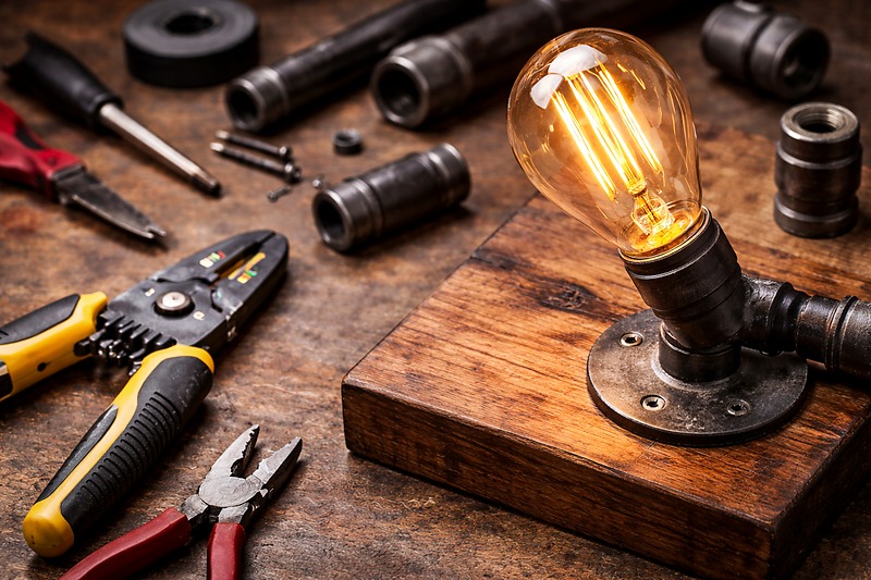 A photo of a workbench covered in tools: wire strippers, black iron pipes, a piece of stained wood, and a glowing amber LED Edison bulb. It looks like a craft project in progress