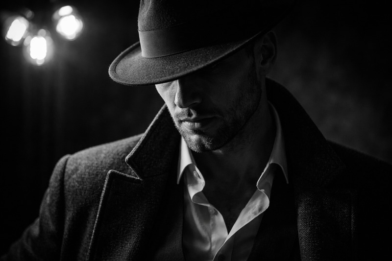 The Moody Key Light A gritty black and white photo of a man wearing a hat. The light is high contrast/chiaroscuro. The light source is a cluster of bare bulbs just out of frame.