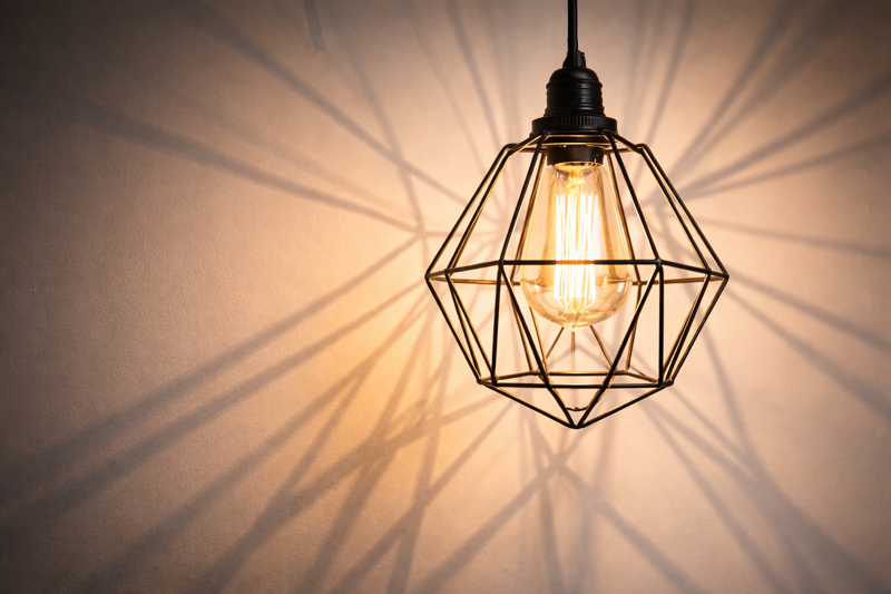 A close up of a black wire cage pendant. The geometric wires cast long, sharp shadow lines on the white wall behind it.